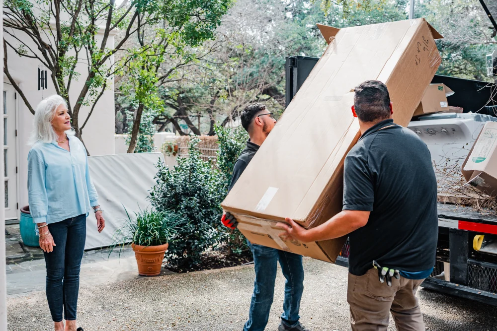 Two movers carry a large cardboard box while a woman observes in a residential setting. TITLE: