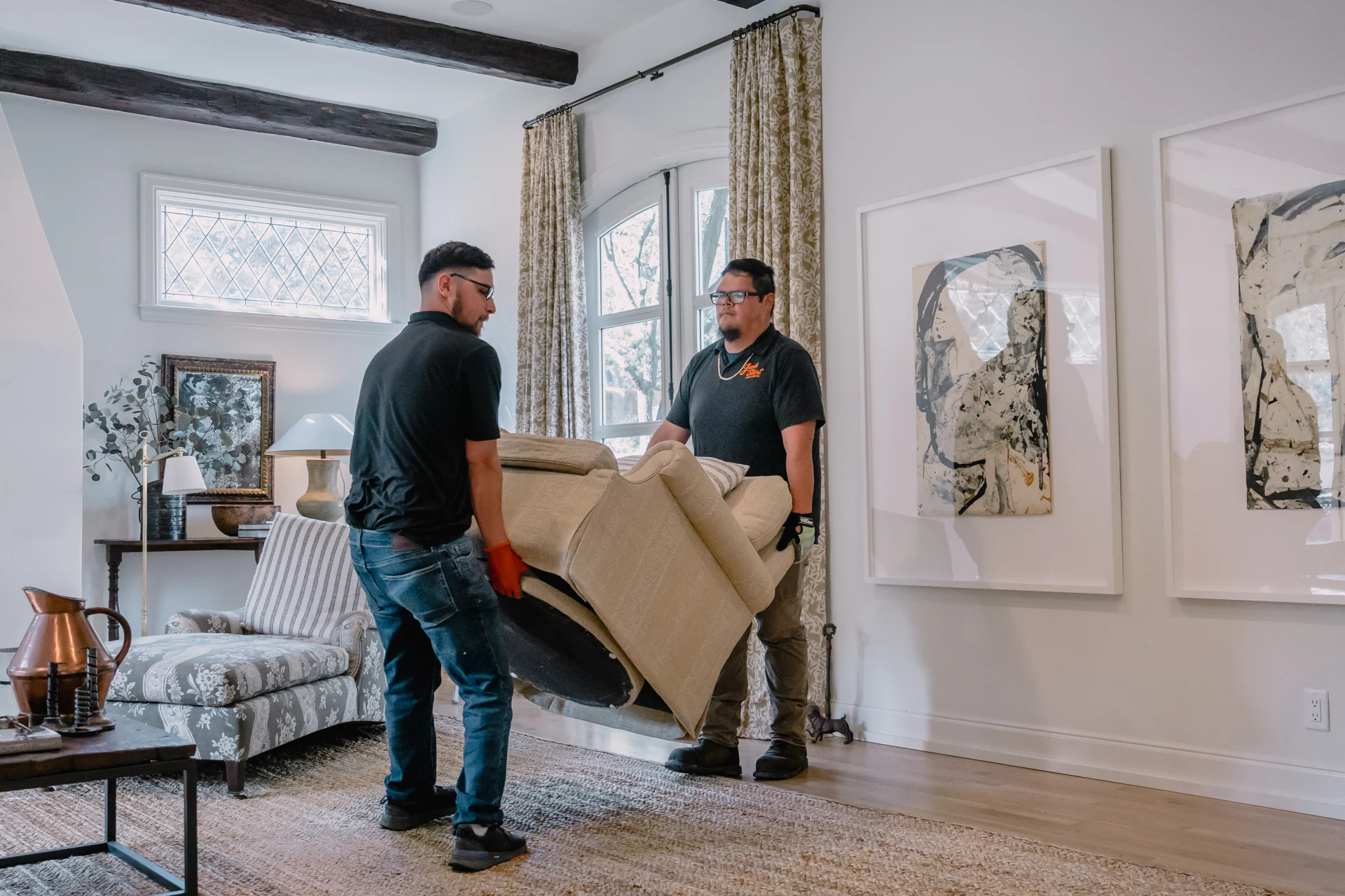 Two professional movers carefully carry a beige armchair through a well-decorated living room. TITLE