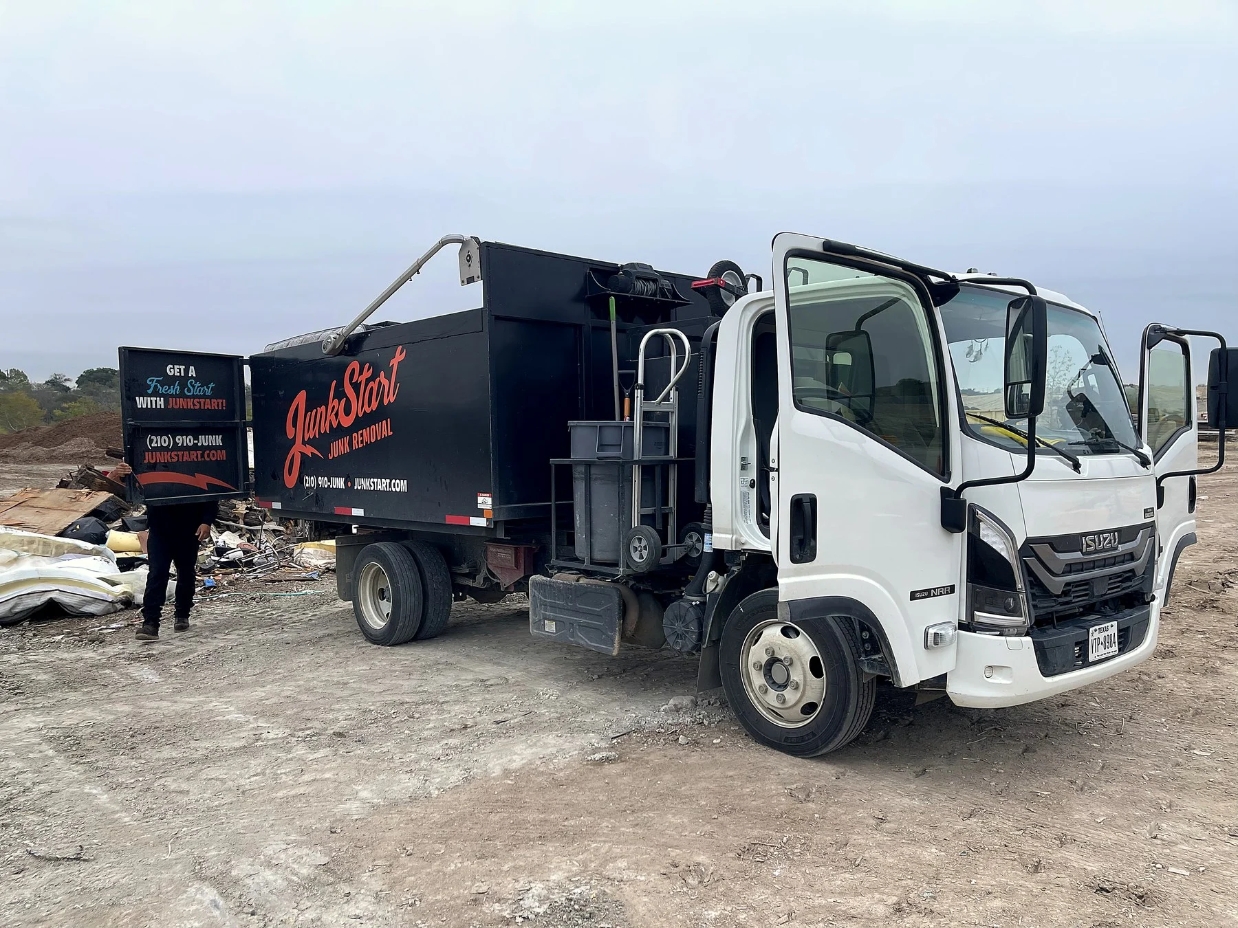 A white JunkStart junk removal truck with open doors is parked on a dirt lot next to a pile of debris.