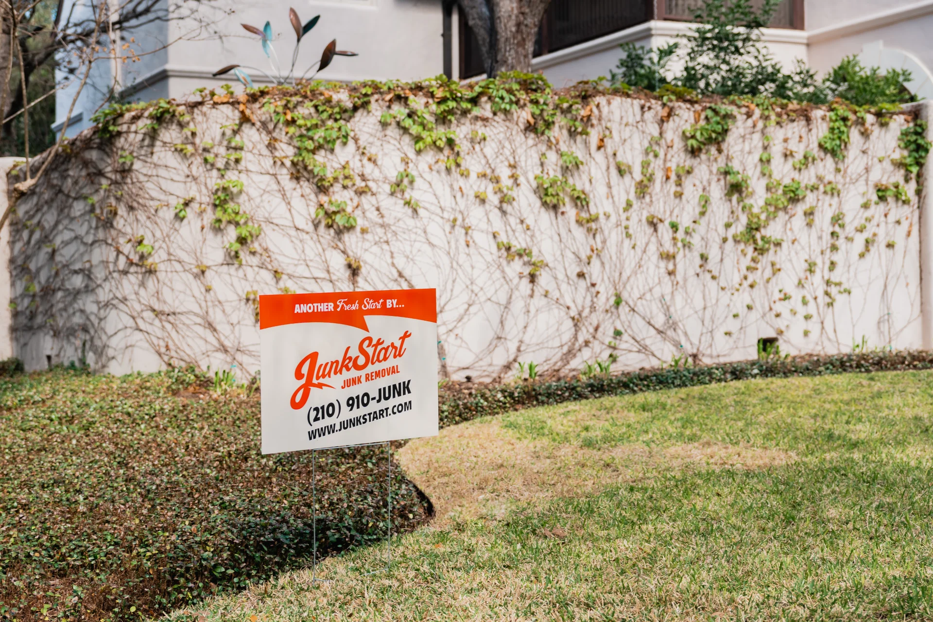 A JunkStart junk removal sign stands in a grassy lawn in front of a vine-