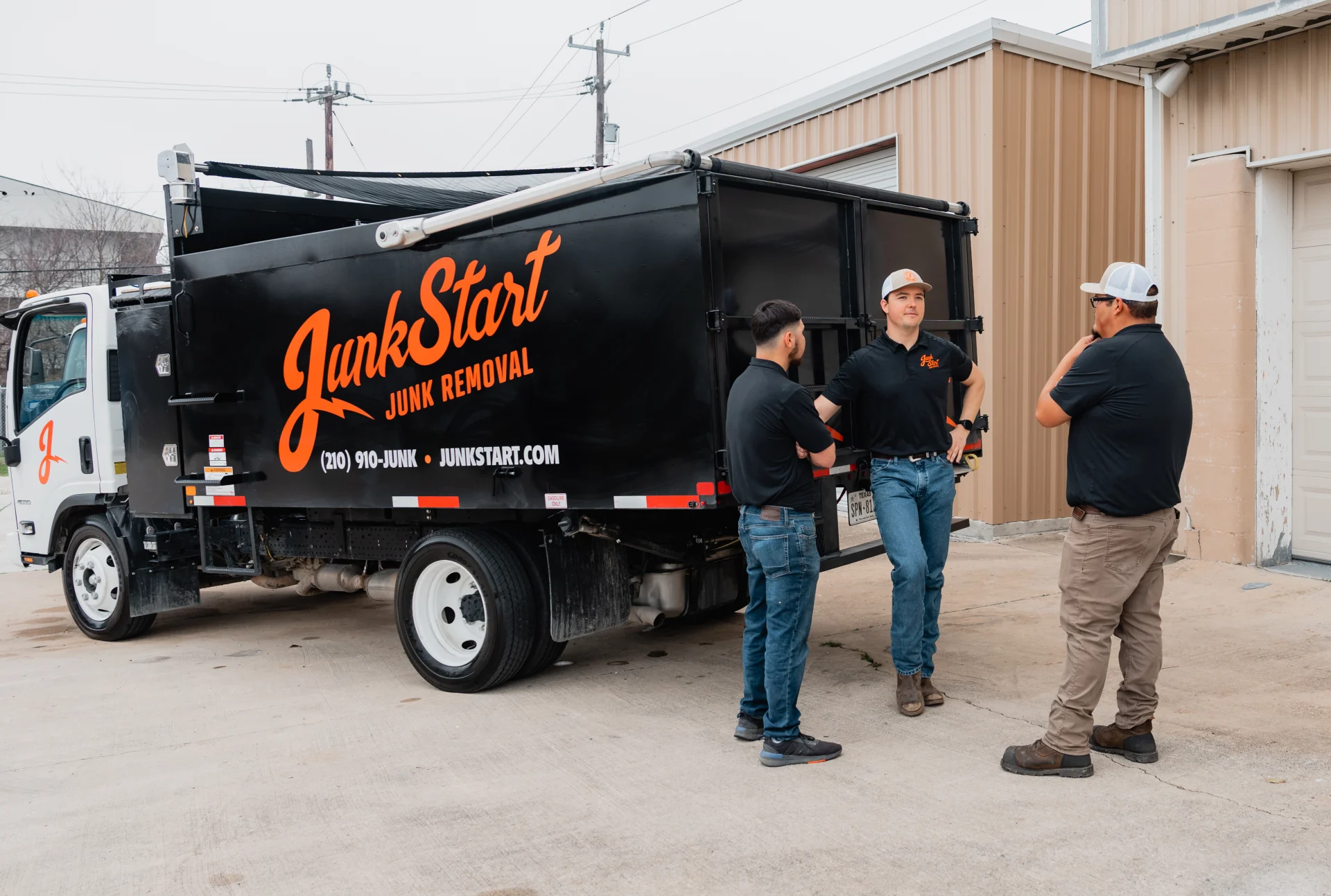 Three JunkStart employees stand beside their black junk removal truck, discussing work outdoors. TITLE