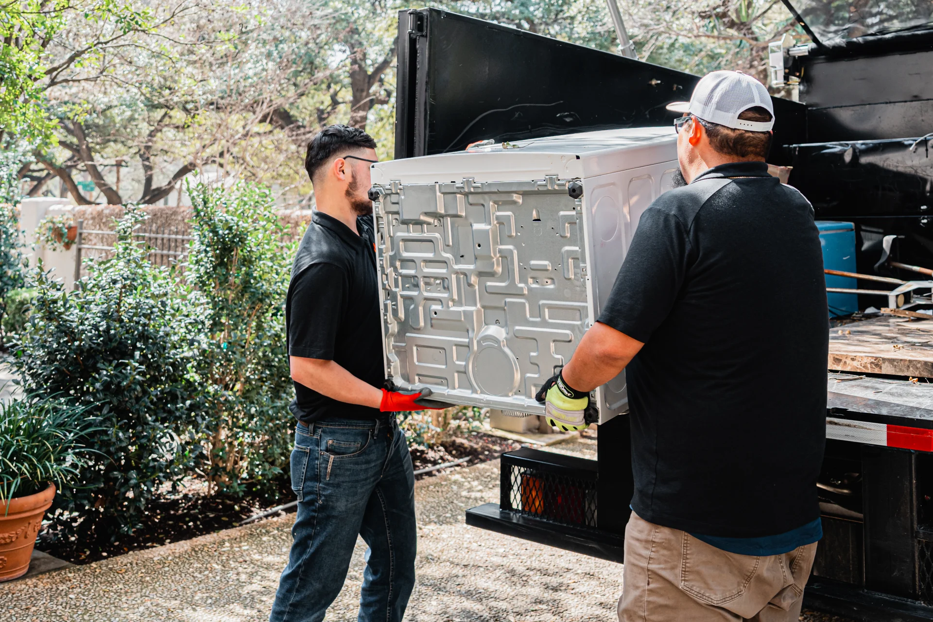 Two workers wearing gloves lift a white appliance onto a truck bed outside.