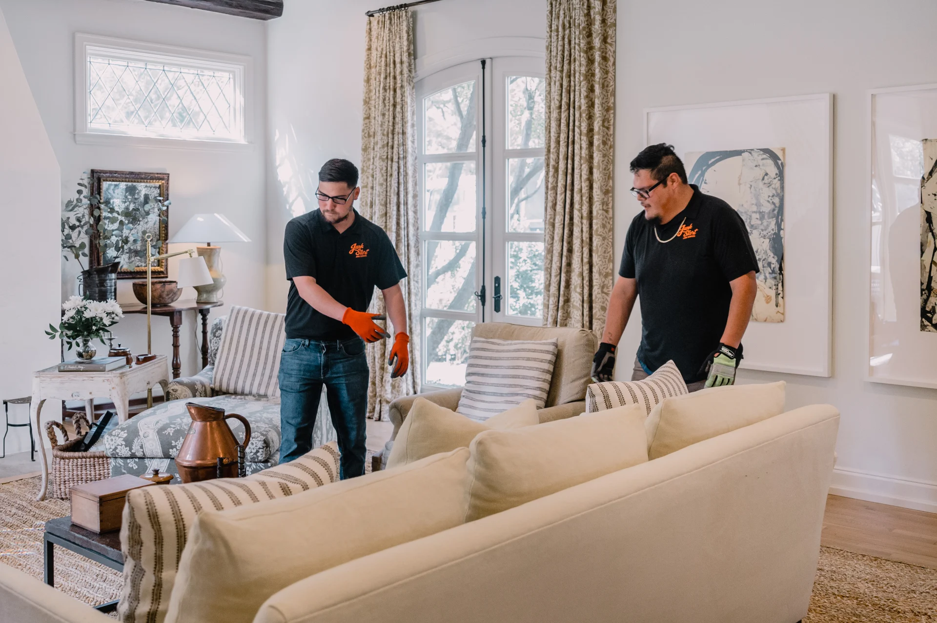 Professional movers in branded shirts and gloves carefully position a light-colored sofa in a stylish living room.