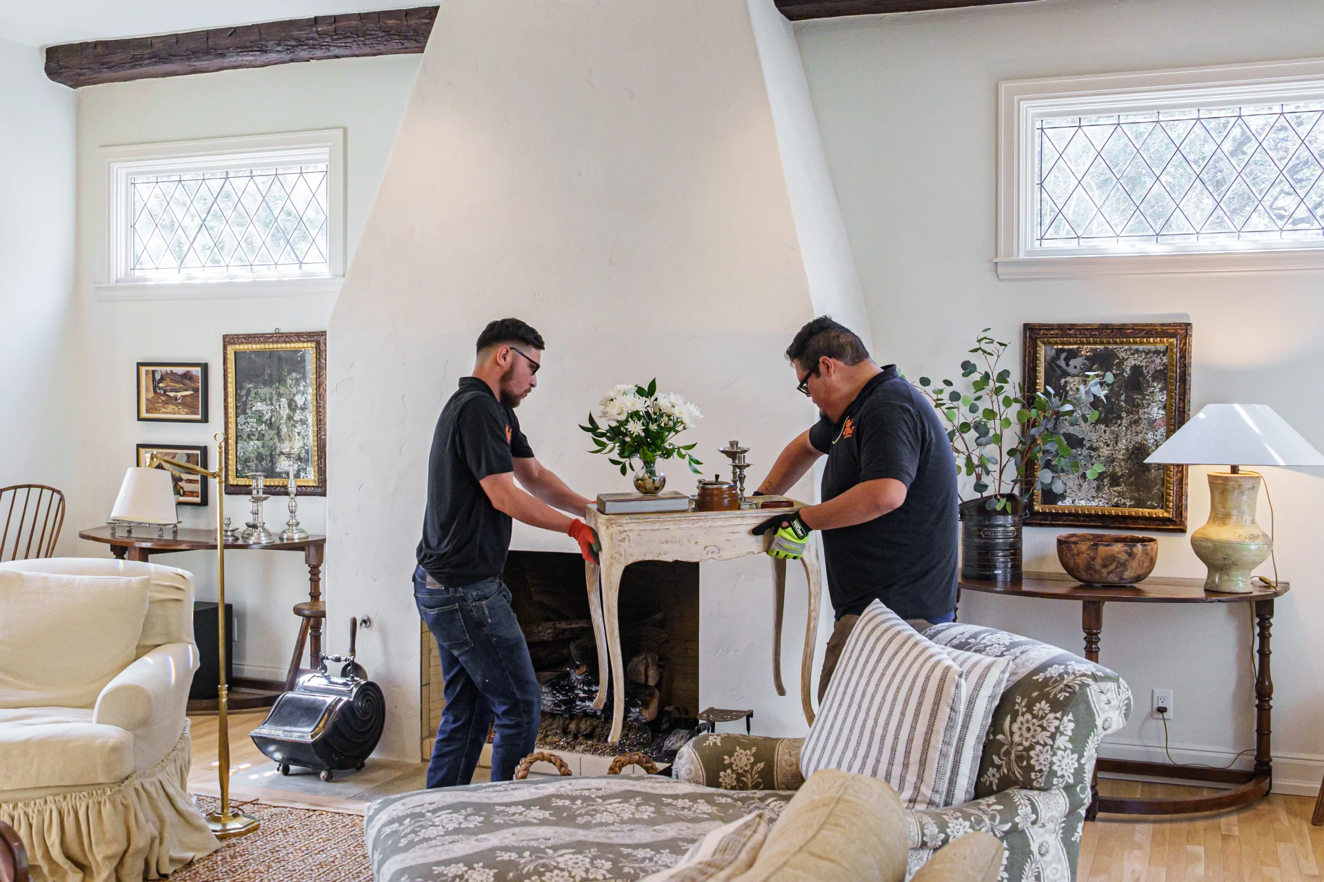 Two men carefully place a decorative table with flowers in a traditional living room.