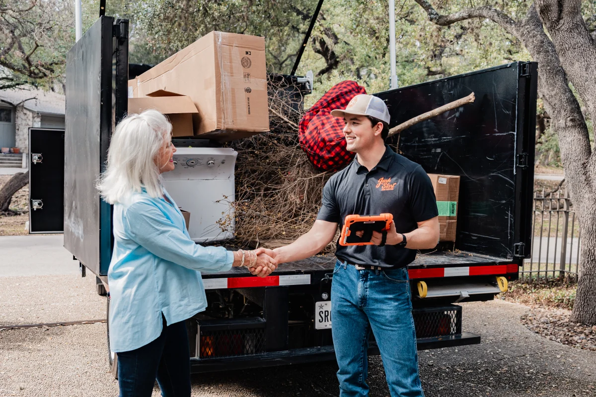 A junk removal employee shakes hands with a happy customer beside a truck filled with debris.